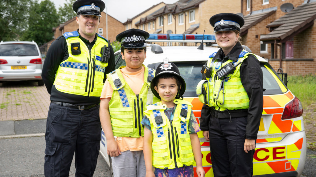 Two police officers with children who are trying on the high vis and hats from the police 