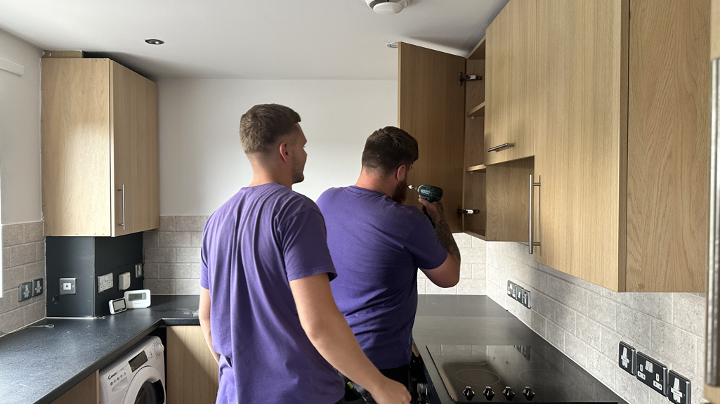 Two people in a kitchen fixing a cabinet door