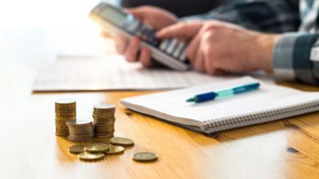 Desk With Money, Pen And Notepad, And Person Using Calculator