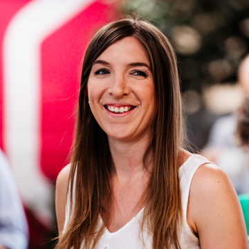 A photo of Sian smiling and looking just off camera. She's got long hair and is wearing a white top with a blurred colourful background. 