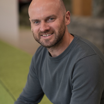 Rob wearing a dark green jumper sitting on an indoor grassy bank and smiling at the camera 