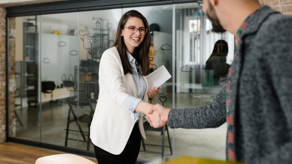 Woman in a white blazer and light blue shirt shaking the hand of a man wearing a more casual grey top. Both are smiling in a modern office.