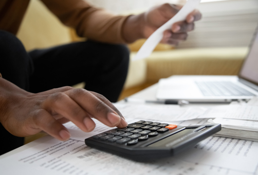 A close up shot of a hand on a calculator on top of some sheets of paper. The person is holding a receipt in the other hand. 