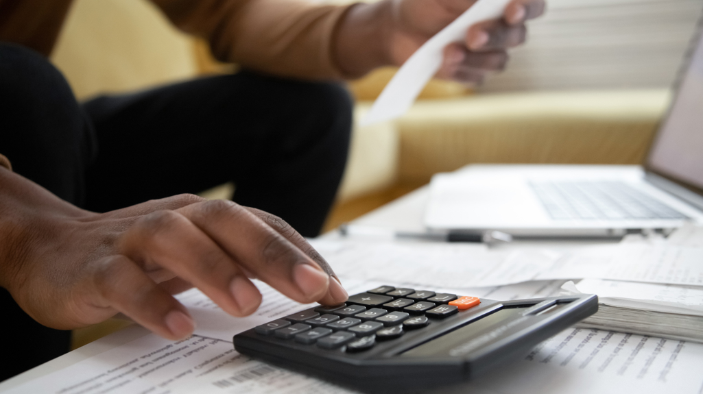 A close up shot of a hand on a calculator on top of some sheets of paper. The person is holding a receipt in the other hand. 