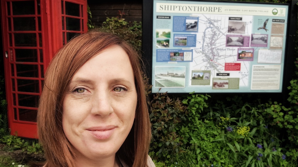 Leanne taking a selfie at Shiptonthorpe outside in front of the town sign with a red phonebook in the background. 