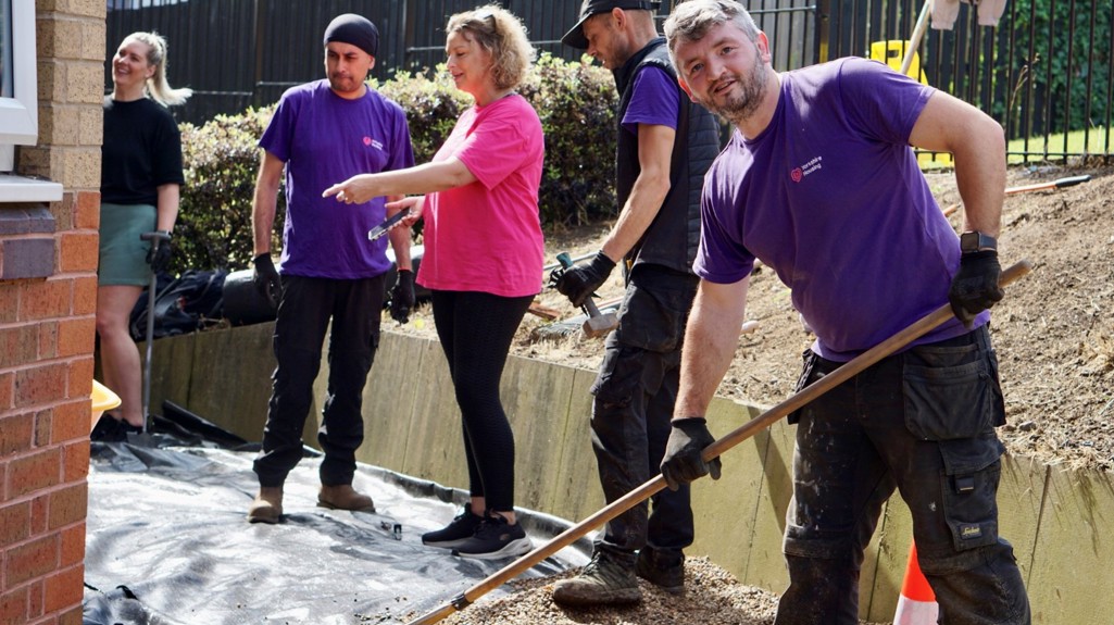 Photo of five people standing alongside a residential building laying grit. The person closest to the camera is looking and smiling at the camera and holding a rake.