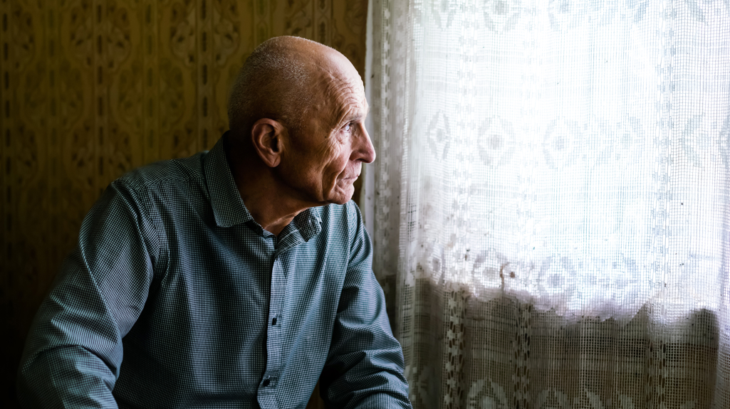 A man looking out the window through a netted curtain. 