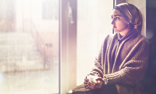 Person sat on a window ledge inside looking out. She's holding a cup of tea and is staring out the window. 