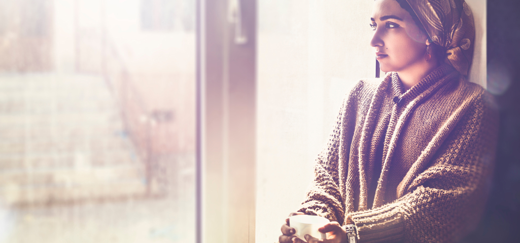 Person sat on a window ledge inside looking out. She's holding a cup of tea and is staring out the window. 