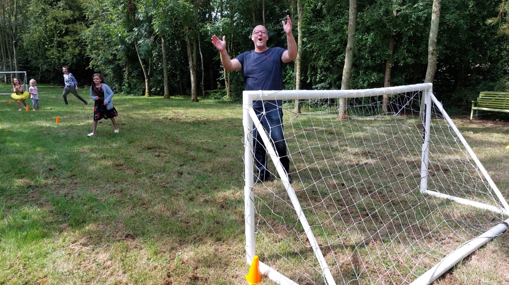 Photo of a grass field lined with trees and a football goal at each end. In between the football goal are 4 children facing the camera and a man in goal looking at the camera with his arms raised.